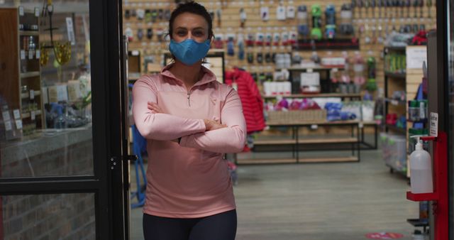 Female store employee or customer wearing a blue face mask and pink sports shirt stands with arms crossed inside a retail store. Many products displayed on shelves in the background. Ideal for use in articles or advertisements about retail businesses, health and safety protocols, pandemic effects on shopping, or indoor activities.