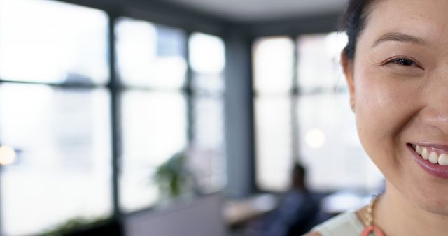 Image showing a close-up of a smiling Asian woman in a modern office. Perfect for articles or websites about workplace happiness, diversity in corporate environments, and the casual professional culture.