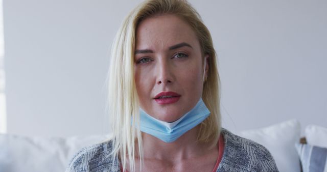 Blonde woman sitting indoors with mask under her chin, looking thoughtful and serious. Useful for topics related to the COVID-19 pandemic, health safety, mental wellness, social issues, and personal reflection.