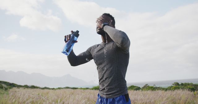 African American man experiencing exhaustion after intense workout in nature, holding water bottle for hydration. Wearing athletic gear, head in hand, standing amidst grassy field with mountains in background, perfect representation of fitness, perseverance, and natural settings. Ideal for health, wellness, fitness promotion, hydration campaigns, athletic wear advertisements.