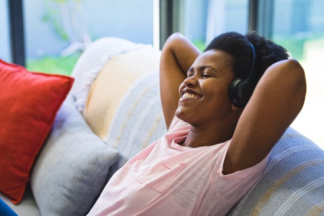 This image depicts a smiling African American mid adult woman enjoying music through headphones while relaxing on a sofa at home. Ideal for use in lifestyle blogs, articles about relaxation and leisure, advertisements for audio equipment, or content promoting mental well-being and self-care.