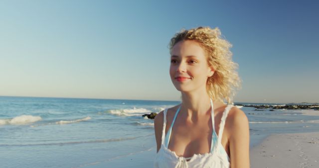 Woman with blonde curly hair walking along sunny beach, smiling and enjoying the moment. Perfect for use in travel and tourism promotions, vacation ads, wellness articles, and lifestyle blogs focusing on relaxation and outdoor activities.