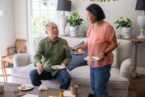 Asian senior couple enjoying cake in cozy living room, woman serving dessert to husband