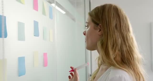 This video depicts a Caucasian woman at work in a creative office setting, looking at memo notes on the wall and pen in hand, suggesting she is in a brainstorming or planning phase. This visual is perfect for illustrating concepts such as teamwork, collaboration, business planning, creative processes, and focus on tasks. It can be used in businessrelated articles, team building workshops, productivity blogs, and motivational posters.