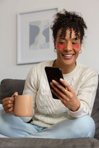 African american woman practicing self-care with hydrogel eye patches and smartphone