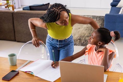 African mother leaning over daughter guiding homework with laptop and smartphone at home