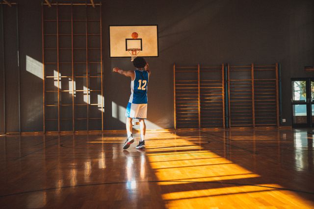 This image captures a basketball player in mid-action, shooting at a hoop in a sunlit gym. Ideal for use in sports-related articles, training guides, motivational posters, and athletic program promotions. The dynamic lighting and composition emphasize the focus and dedication of the athlete.