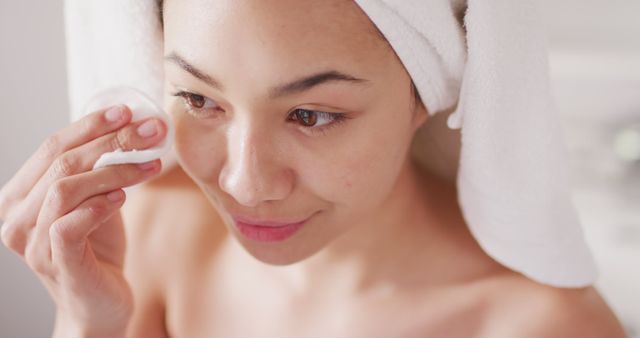 Woman cleansing her facial skin with a cotton pad in bathroom. Suitable for articles and advertisements about skincare routines, beauty tips, self-care, hygiene, and wellness products.