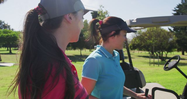 Women golfers stand beside a golf cart on a sunny day, engaging in a discussion focused on their game strategy. The sun shines brightly, creating a vibrant and active atmosphere typical for a sports field. This image is ideal for content related to women in sports, outdoor activities, golf events, teamwork, and promoting female athletes.