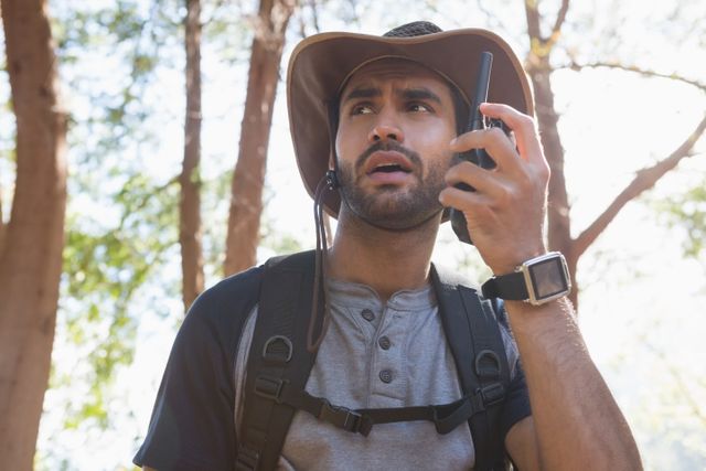 Man using walkie-talkie in forest on sunny day, ideal for themes of outdoor adventure, communication, and safety. Suitable for articles, blogs, and advertisements related to hiking, technology in nature, and wilderness exploration.