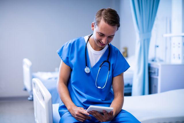 Young doctor in blue scrubs sitting on hospital bed using digital tablet. Ideal for illustrating modern healthcare, medical technology, and professional healthcare settings. Useful for medical websites, healthcare blogs, and educational materials.