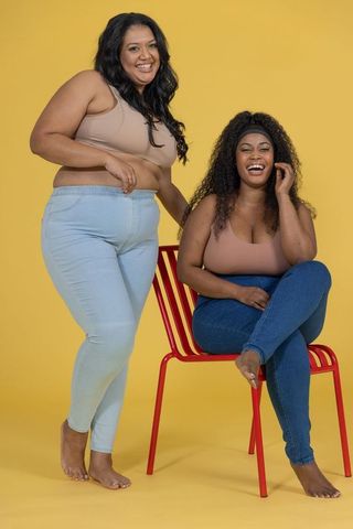 Curvy friends laughing and posing in jeans and tank tops on bright yellow backdrop