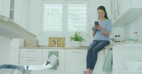 Relaxed Asian Woman Enjoying Smartphone and Coffee in Modern Kitchen