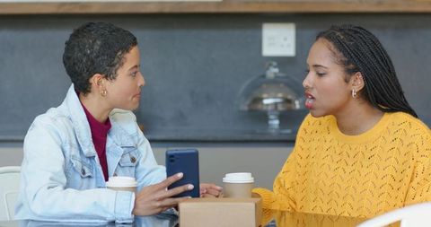 Two african american women sharing coffee and discussing smartphone at cafe table