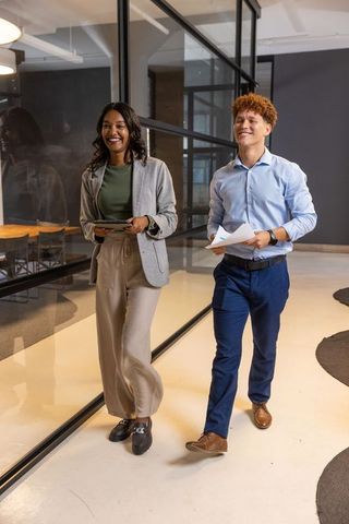 Smiling diverse coworkers walking in modern office corridor holding tablet and documents