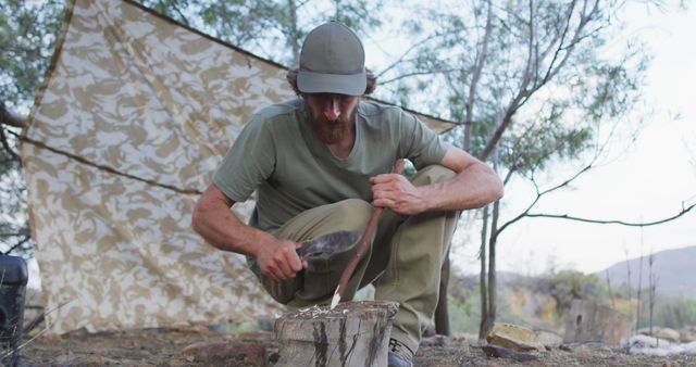 Bearded caucasian male survivalist sharpening stick with machete at camp in wilderness. exploration, travel and adventure, survivalist in nature.