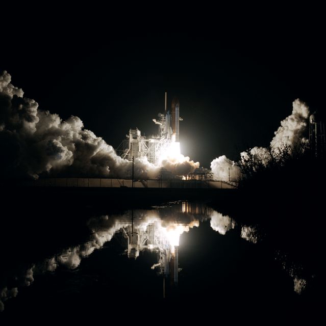 Photo captures nighttime launch of Space Shuttle Columbia from Kennedy Space Center dated January 12, 1986. The vibrant scene showcases powerful liftoff and smoke clouds, beautifully reflected in water. Suitable for educational materials about NASA, space exploration, historical missions, or presentations on technological advancements in space travel.