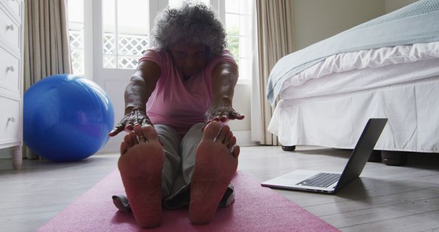 Senior woman engaging in yoga practice on a pink exercise mat, following potentially a tutorial on her laptop. Image suitable for health and fitness websites, promoting home workouts, wellness programs for seniors, or online fitness classes.