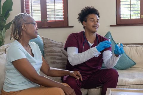 Home nurse preparing vaccine injection while senior african american woman holding knee