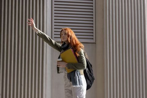 Redhead student hailing ride outside modern campus columns holding coffee and notebooks