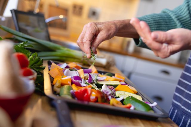 This image depicts a senior woman in a kitchen preparing a healthy vegetable dish. She is sprinkling herbs on chopped vegetables while using a tablet. This image can be used for articles or advertisements related to healthy eating, senior lifestyle, home cooking, nutrition, and culinary activities.