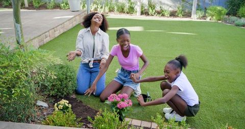 Joyful family gardening together: mother and daughters planting pink flowers in sunny yard
