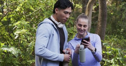 Active diverse couple checking fitness app while holding water bottles on wooded trail