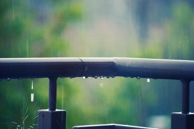 Water droplets forming on metal balcony railing on rainy day. Background is blurred with lush greenery and gentle downpour. Perfect for projects related to weather, nature, rainy days, and outdoor lifestyle.