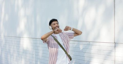 Young indian man listening to music and dancing with headphones on sunlit brick wall