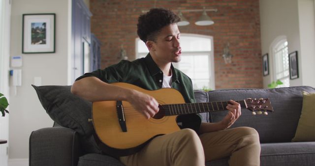 Young man in casual attire playing acoustic guitar and singing on living room sofa. Ideal for use in lifestyle blogs, music websites, home decor magazines, and advertisements promoting relaxation or leisure activities at home.
