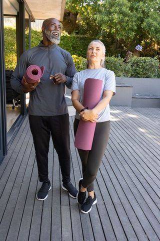 Diverse couple holding yoga mats standing on wooden deck at modern home practicing wellness