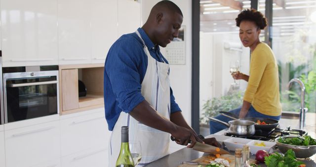 Couple cooking together in a stylish, modern kitchen. Man chopping vegetables on a cutting board while woman drinks wine and looks on with a smile. Stainless steel appliances and bright natural light create an inviting atmosphere. Ideal for websites and advertisements focused on home life, cooking, modern living, and relationship activities.