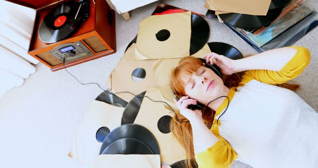 A young woman with orange hair is lying on a carpeted floor, surrounded by vinyl records, wearing casual attire and headphones. She is enjoying the music, fully immersed in the moment. This scene is perfect for illustrating leisure and relaxation at home, vintage music elements, or cozy domestic vibes.