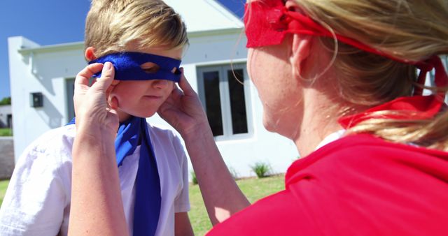 A Caucasian woman is adjusting a blue mask on a young boy's face, both wearing superhero capes, with copy space. Their playful attire and actions suggest a fun, imaginative playtime activity outdoors.