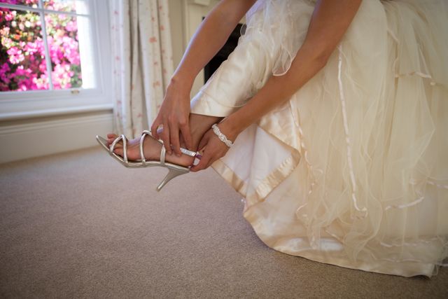 This image captures a bride adjusting her sandals while wearing a wedding dress at home. It is perfect for use in wedding blogs, bridal magazines, and advertisements for bridal accessories or wedding services. The focus on the bride's lower section and the elegant details of her dress and sandals highlight the preparation and anticipation of the wedding day.