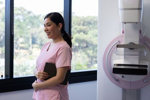 Smiling Nurse in Pink Scrubs Holding Tablet beside Mammography Unit for Breast Care