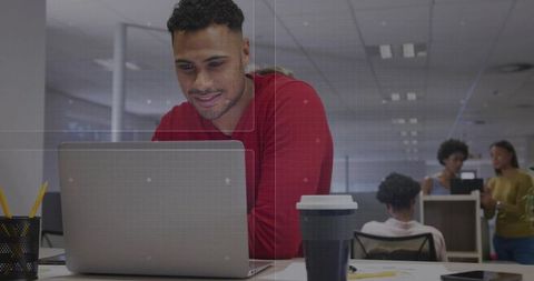 Man leaning on laptop in open-plan office collaborating with team, reusable coffee cup