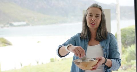 Mature woman stirring wooden bowl in coastal kitchen overlooking bay and horizon