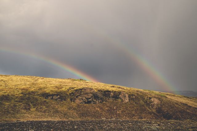 Double rainbow arches over a grassy hill under an overcast sky. This image is ideal for use in weather-related content, environmental articles, nature-themed media, and backgrounds. Showcases natural beauty and atmospheric phenomena in the countryside.
