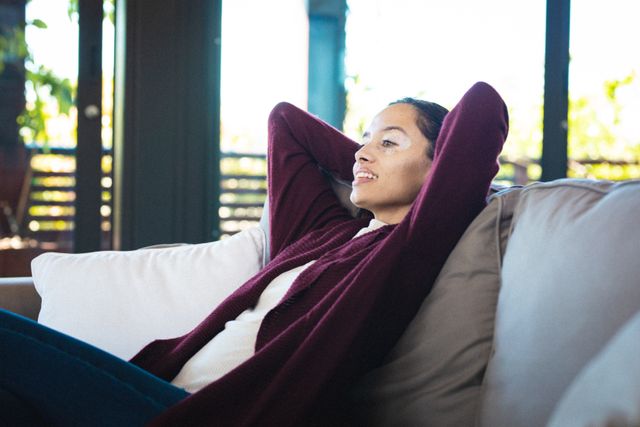 This image depicts a biracial woman with vitiligo relaxing on a sofa at home, smiling with her hands behind her head. The natural light and comfortable setting make it ideal for use in lifestyle blogs, articles on relaxation and self-care, or advertisements promoting home comfort and leisure activities.