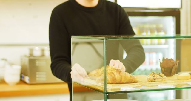 Barista arranging fresh pastries in bakery display case, includes croissant and muffin. Ideal for use in marketing materials for cafes or bakeries, food and beverage promotions, or small business advertisements focusing on fresh products and customer service.