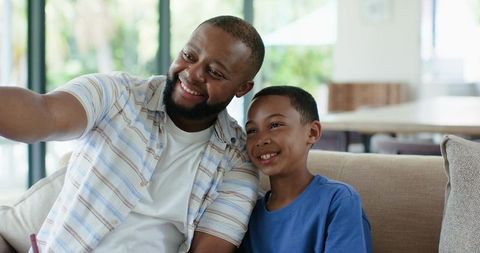 African american father and son taking selfie on sunlit living room sofa, smiling and bonding