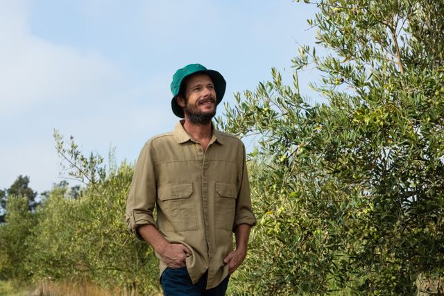 Man standing in an olive grove, smiling and looking content. Wearing a green hat and casual clothing, with hands in pockets. Ideal for use in agricultural promotions, rural lifestyle blogs, or nature-related content.