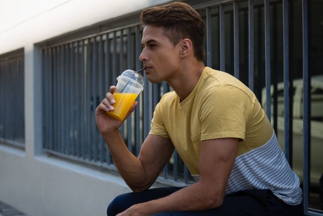 Young man enjoying a refreshing juice while sitting by a wall in an urban environment. Ideal for use in advertisements promoting healthy lifestyles, summer activities, or urban living. Can also be used in blogs or articles about city life, fitness, or casual fashion.