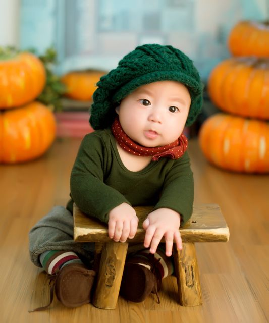 Adorable baby wearing green beanie and red scarf, sitting on small wooden bench with pumpkins in background. Ideal for autumn-themed promotions, family-oriented advertisements, baby clothing catalogs, or seasonal social media posts. Highlights innocence and warmth, perfect for seasonal promotions and festive events.