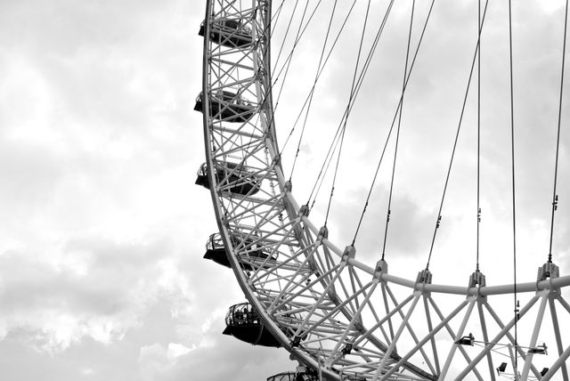 Partial view of Ferris wheel in grayscale against a cloudy sky. Ideal for use in travel brochures emphasizing moody vintage aesthetic, educational materials about London landmarks, and blog posts relating to travel or city life.