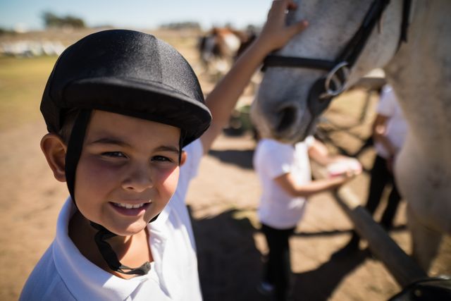 Young boy wearing helmet smiling while touching a white horse at an equestrian center. Ideal for use in advertisements for equestrian activities, children's outdoor activities, summer camps, and animal bonding experiences.