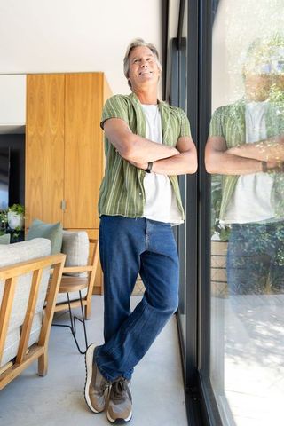 Mature man leaning by glass door in modern sunlit living room, relaxed casual lifestyle