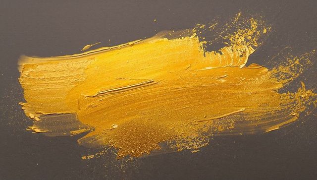 Metallic gold paint stroke on dark gray background with textured impasto sheen