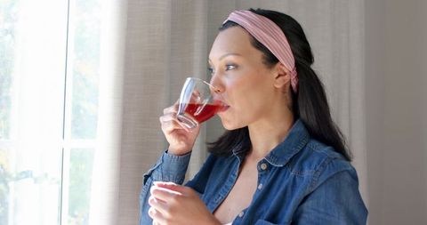Mid adult woman in denim sipping beverage and reflecting at home window